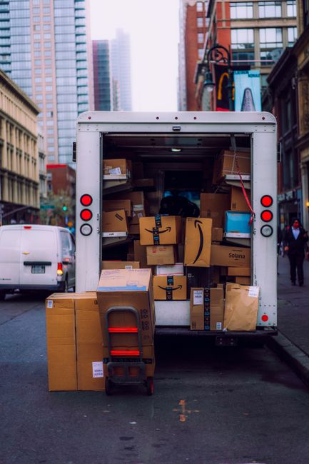 A man wearing a white T-shirt and jeans is stacking cardboard boxes outside a white delivery van parked on the street near a residential property with a white house, a black roof, and a wooden fence.