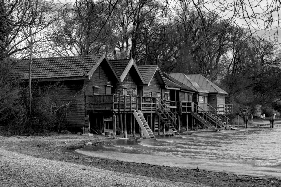 A row of wooden beach huts elevated on stilts along the shoreline of a body of water, with wooden stairs leading from the beach to each hut's small porch. The huts have pitched roofs with shingle cove